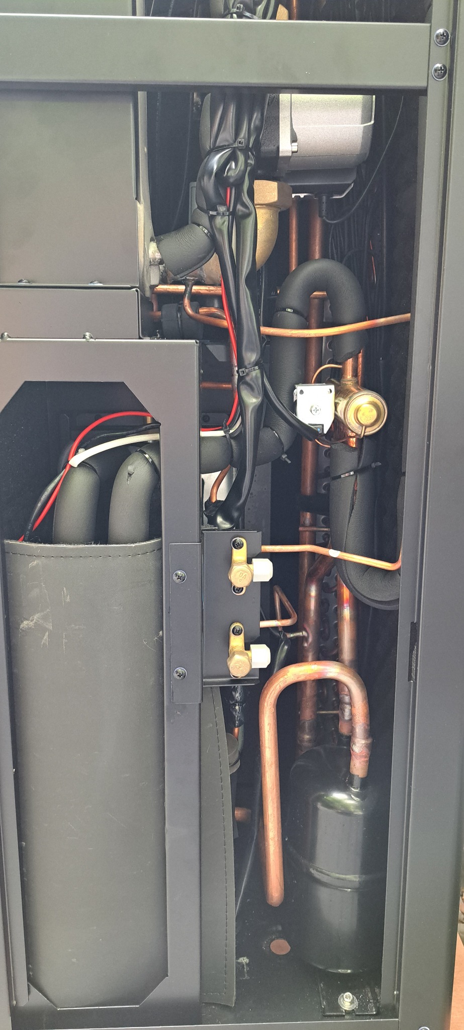Wnętrze czarnej obudowy z miedzianymi rurami, zaworami i przewodami elektrycznymi, prawdopodobnie element instalacji grzewczej lub chłodniczej.