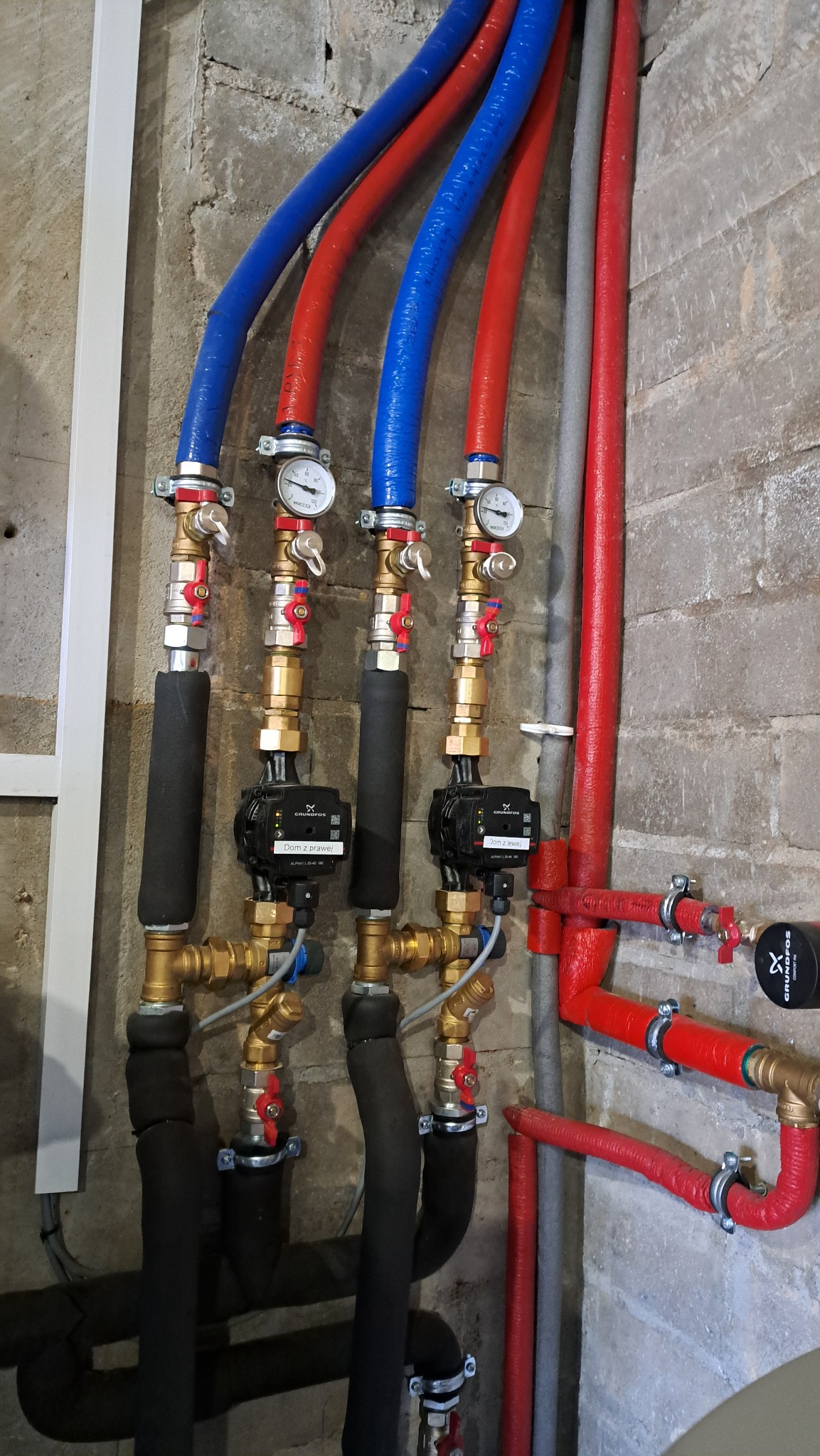 Instalacja hydrauliczna z rurami w kolorze niebieskim i czerwonym, wyposażona w pompy obiegowe Grundfos z etykietami 'Dom z prawej' i 'Dom z lewej', manometry i zawory, zamontowana na surowej...