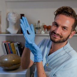 Magic Therapies Gabinet Medycyny Naturalnej - Mężczyzna w niebieskim uniformie i rękawiczkach prezentuje pijawkę na dłoniach, w tle gabinet z regałem, na którym stoją książki i dekoracje.