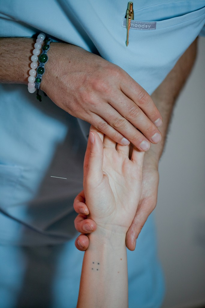 Cienka igła wkłuta w dłoń pacjenta, widoczne dłonie terapeuty w niebieskim uniformie z piórem w kieszeni, bransoletki na nadgarstku terapeuty i mały tatuaż na nadgarstku pacjenta.