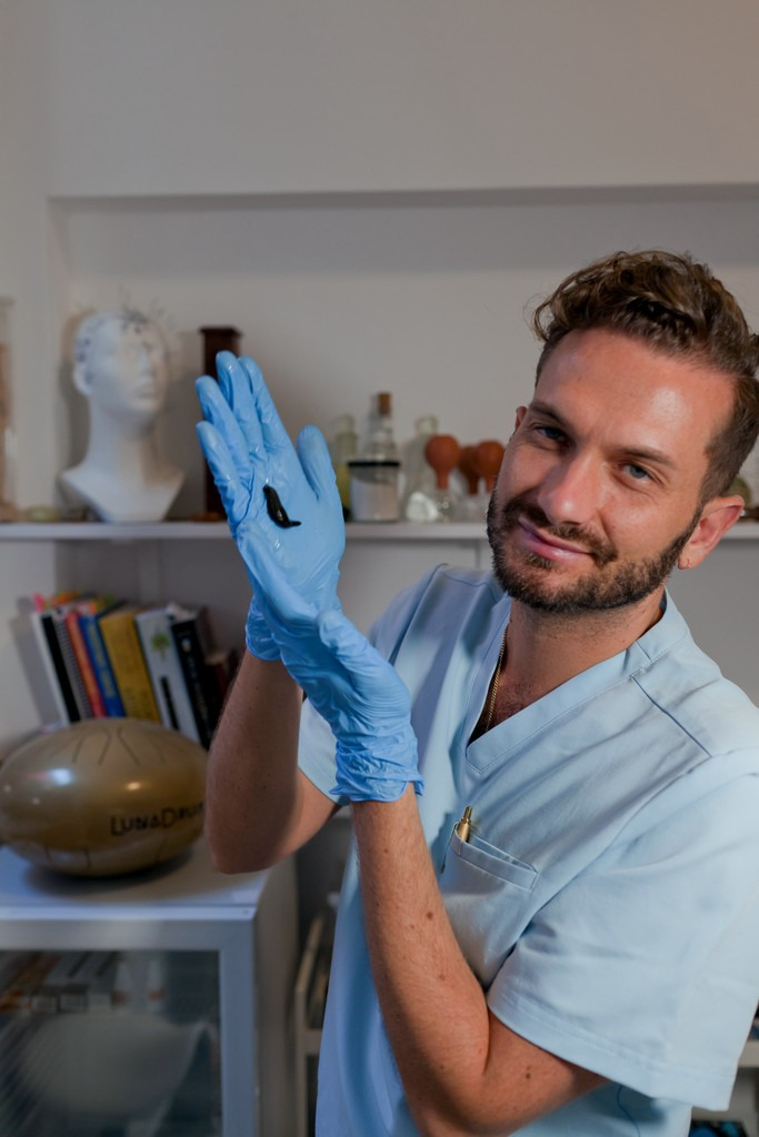 Mężczyzna w niebieskim uniformie i rękawiczkach prezentuje pijawkę na dłoniach, w tle gabinet z regałem, na którym stoją książki i dekoracje.
