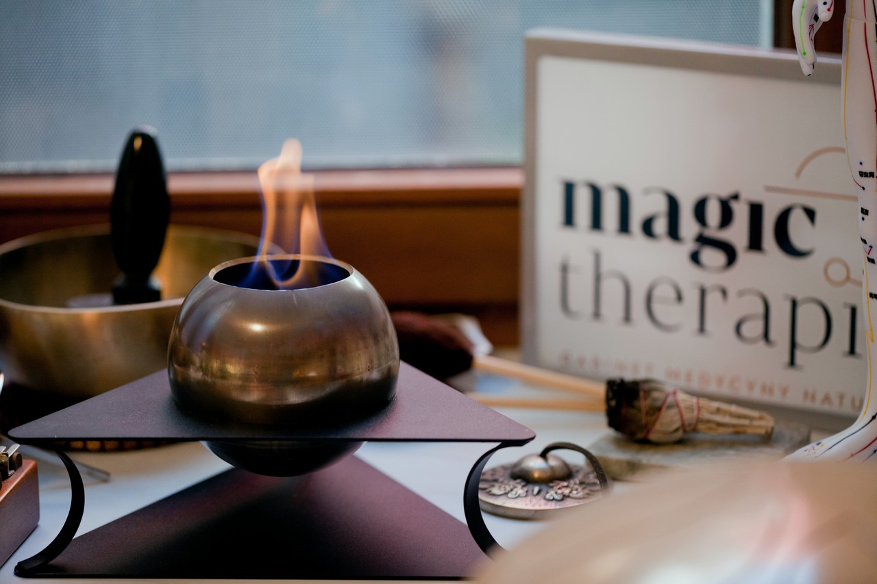 Metalowa miseczka z płomieniem wewnątrz, ustawiona na czarnym trójkątnym stojaku, obok misy tybetańskiej i modelu anatomicznego z punktami akupunkturowymi, w gabinecie w Krakowie. W tle widoczny...