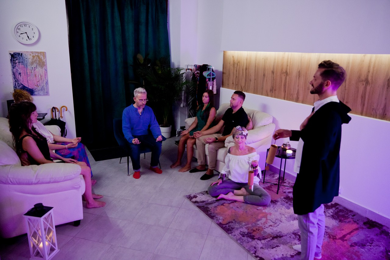 Grupa osób w trakcie sesji relaksacyjnej w pomieszczeniu z fioletowym oświetleniem, prowadzący z instrumentem perkusyjnym stoi przed siedzącymi.
