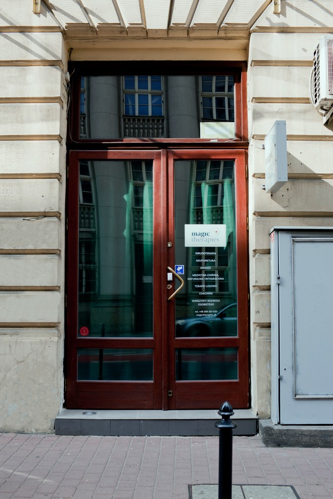 Wejściowe, dwuskrzydłowe, przeszklone drzwi do gabinetu 'magic therapies' z listą oferowanych usług: hirudoterapia, akupunktura, masaż, medycyna chińska, terapia ludowa, coaching, warsztaty rozwoju...