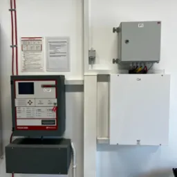 Centrala alarmowa Polon 4200 na ścianie, z instrukcją i schematem. Widoczne okablowanie i skrzynki elektryczne. Elementy systemu alarmowego wewnątrz budynku.