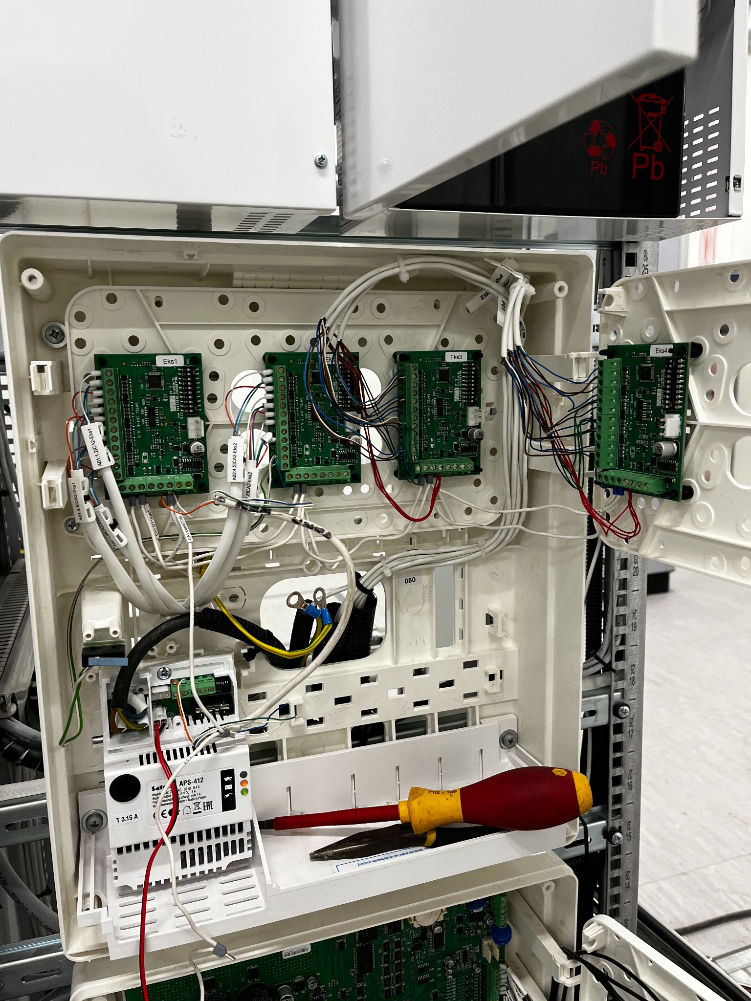 Otwarta obudowa centrali alarmowej z modułami EKS1, EKS2, EKS3, EKS4, przewodami i zasilaczem APS-412 Satel. Widoczny śrubokręt. Element instalacji alarmowej.