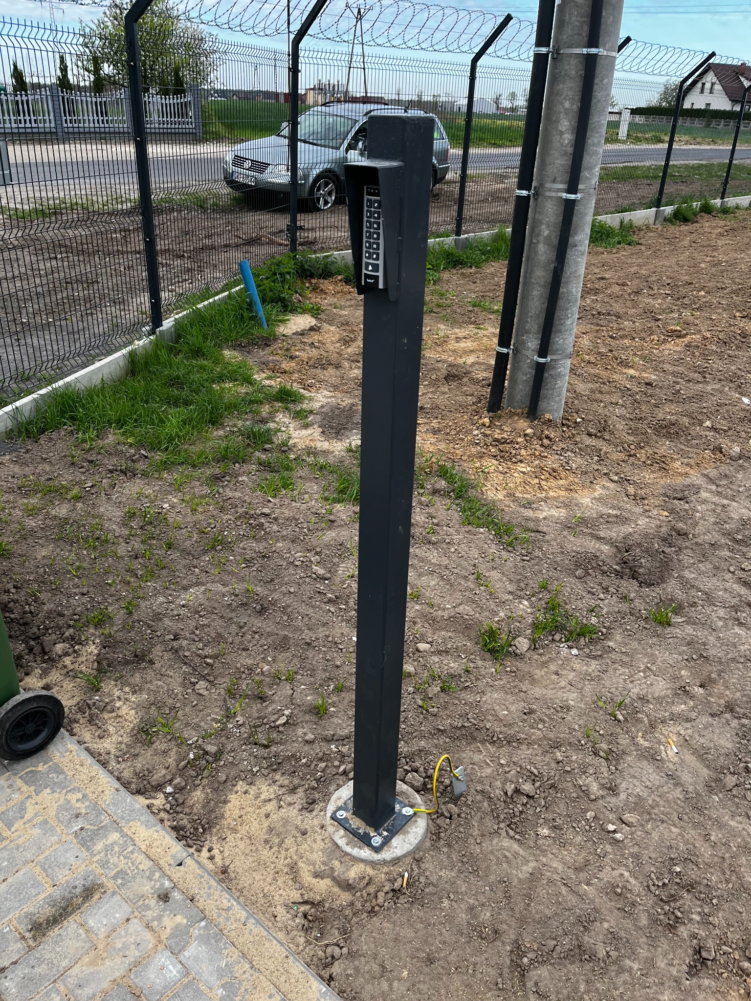 Zewnętrzny panel dostępu z klawiaturą numeryczną zamontowany na słupku przy ogrodzeniu z drutem kolczastym. Widoczny samochód i słupy z przewodami.