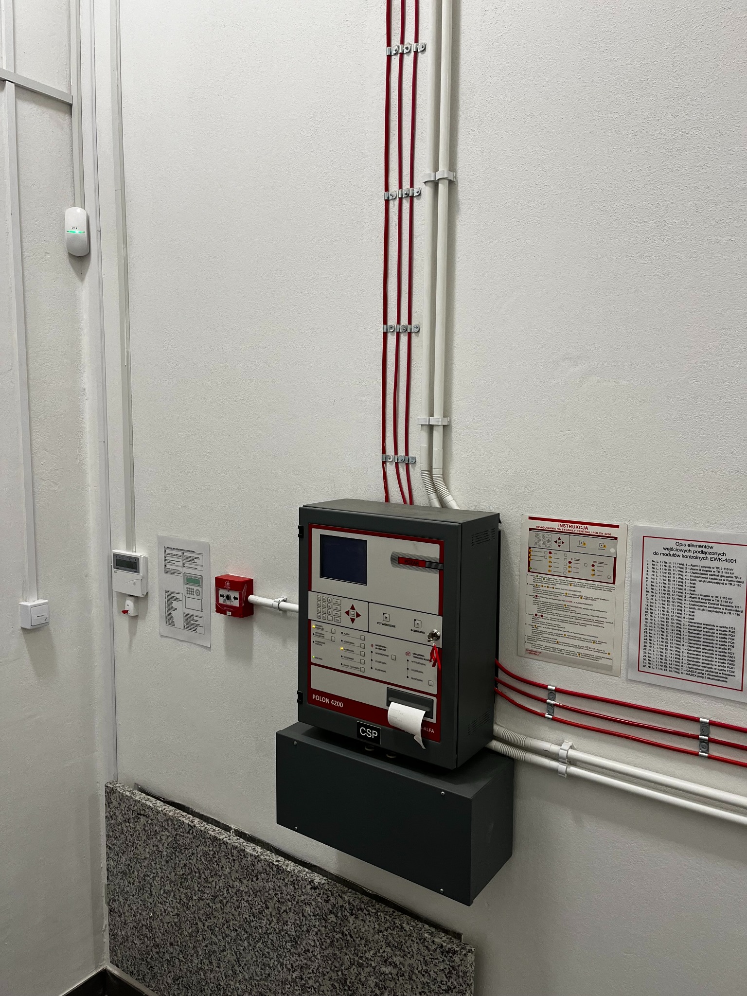 Centrala alarmowa Polon 4200 zainstalowana na ścianie, z widocznymi kablami w rurkach i czujnikiem dymu. Element systemu alarmowego.