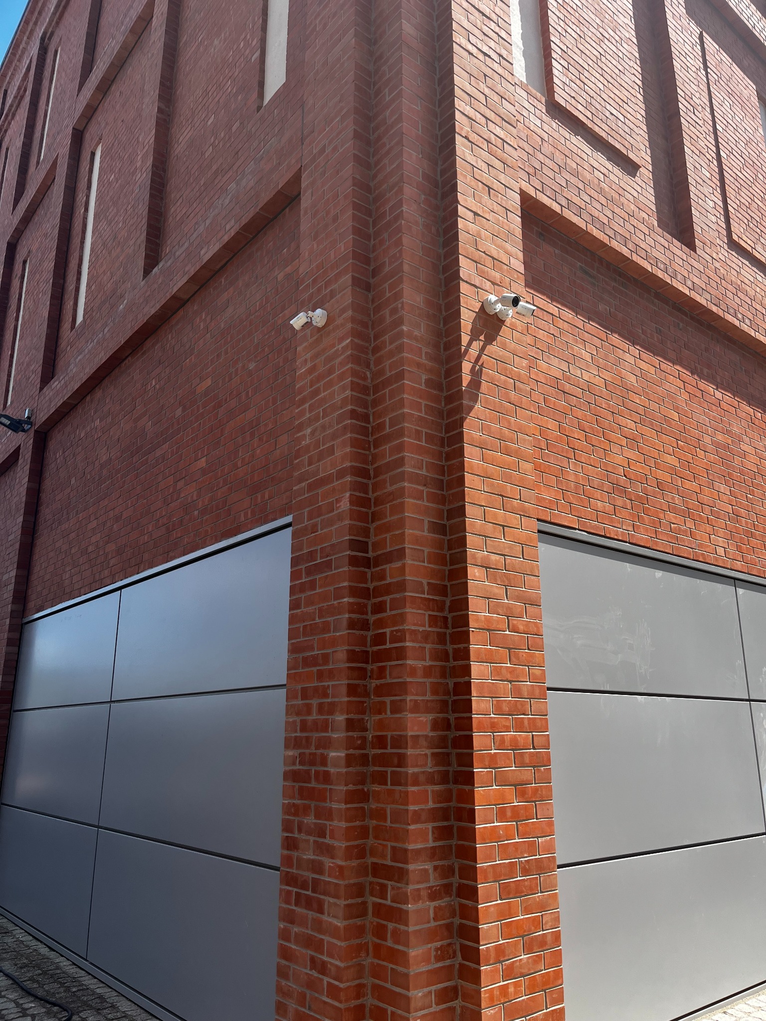 Dwie kamery monitoringu na rogu budynku z czerwonej cegły i szarych paneli elewacyjnych. Widok z dołu, oświetlenie dzienne.