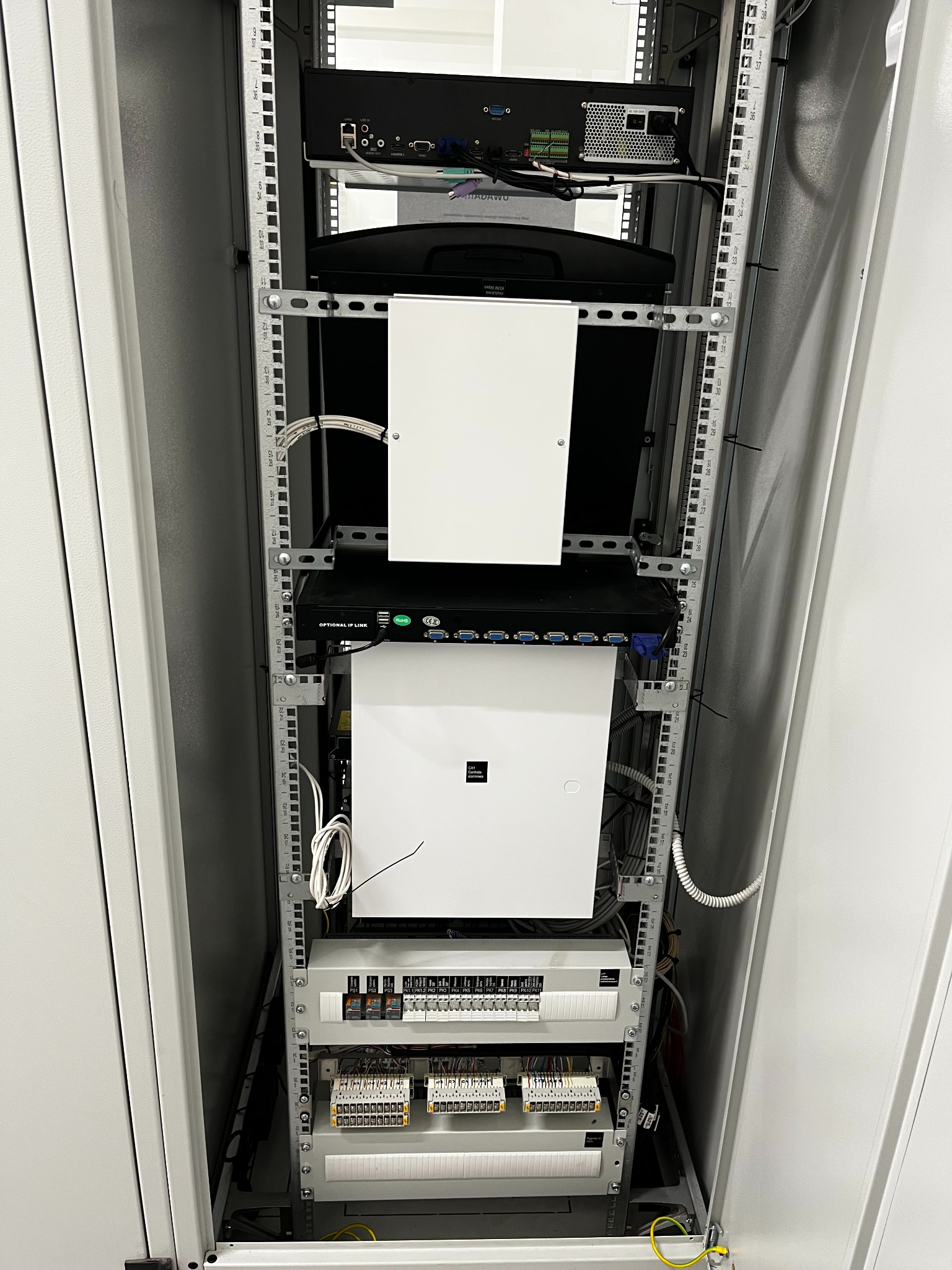 Szafa rack z zamontowanymi urządzeniami: rejestrator, monitor, router i zasilacz. Widoczne przewody i listwy zaciskowe. Rozwiązanie dla monitoringu.