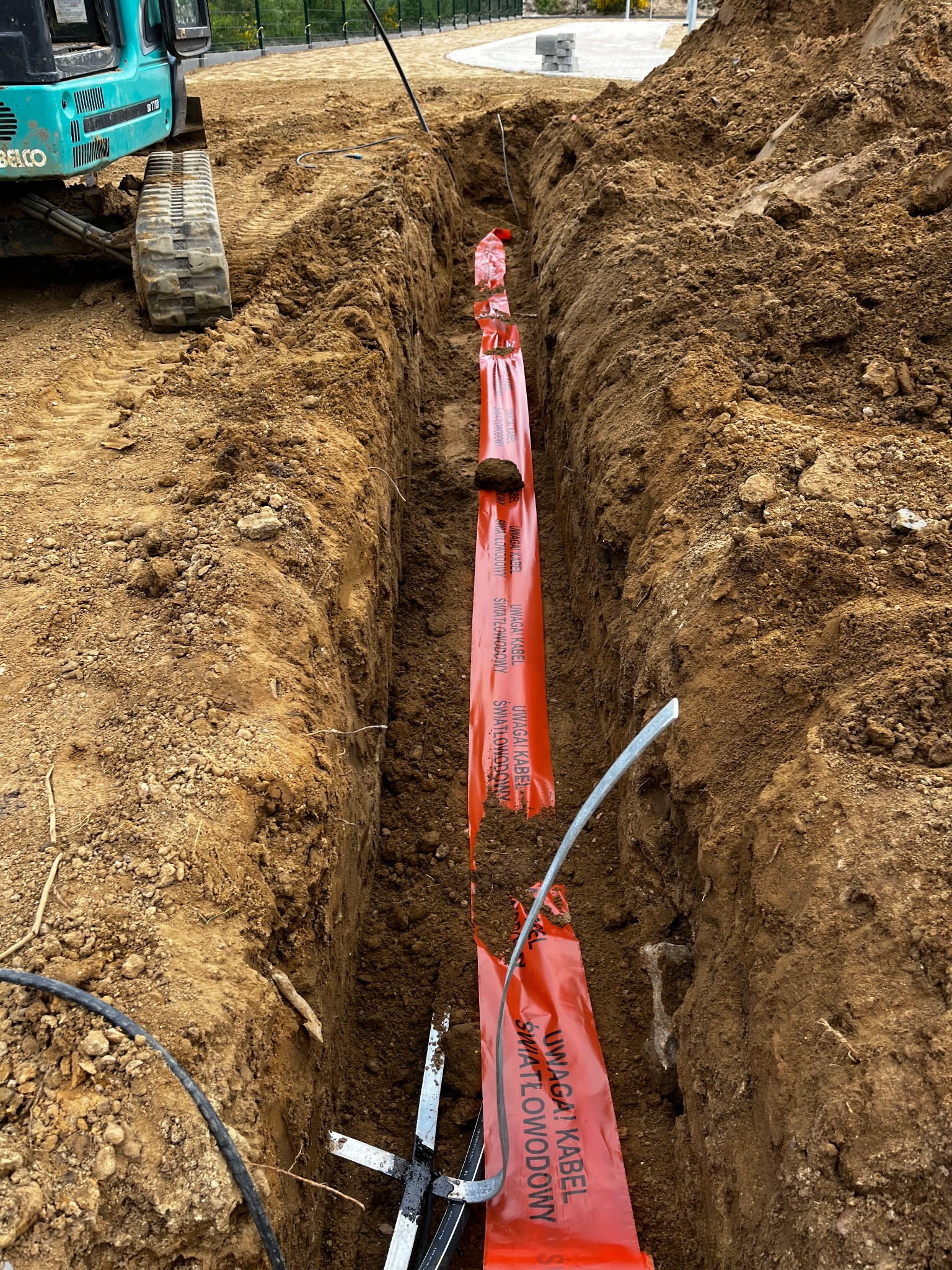 Wykop z pomarańczową taśmą ostrzegawczą 'Uwaga! Kabel światłowodowy' wzdłuż wykopu, obok koparka. Etap prac instalacyjnych kabla światłowodowego.