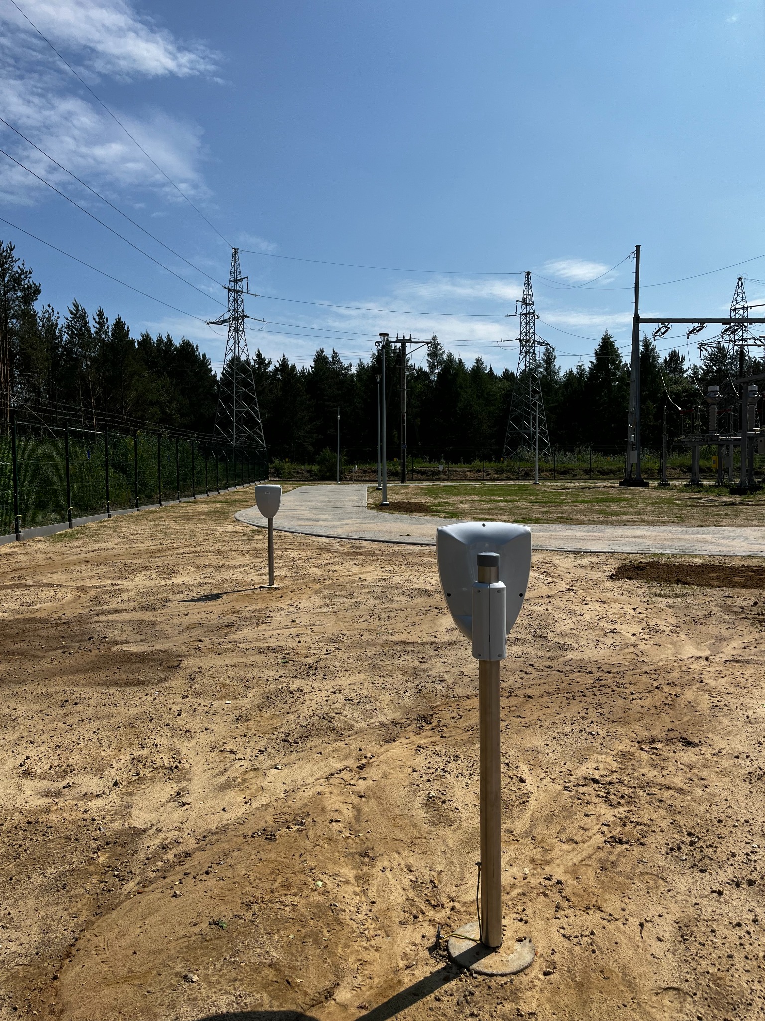 Dwa białe głośniki tubowe na słupkach na terenie stacji transformatorowej, w tle słupy wysokiego napięcia i las.