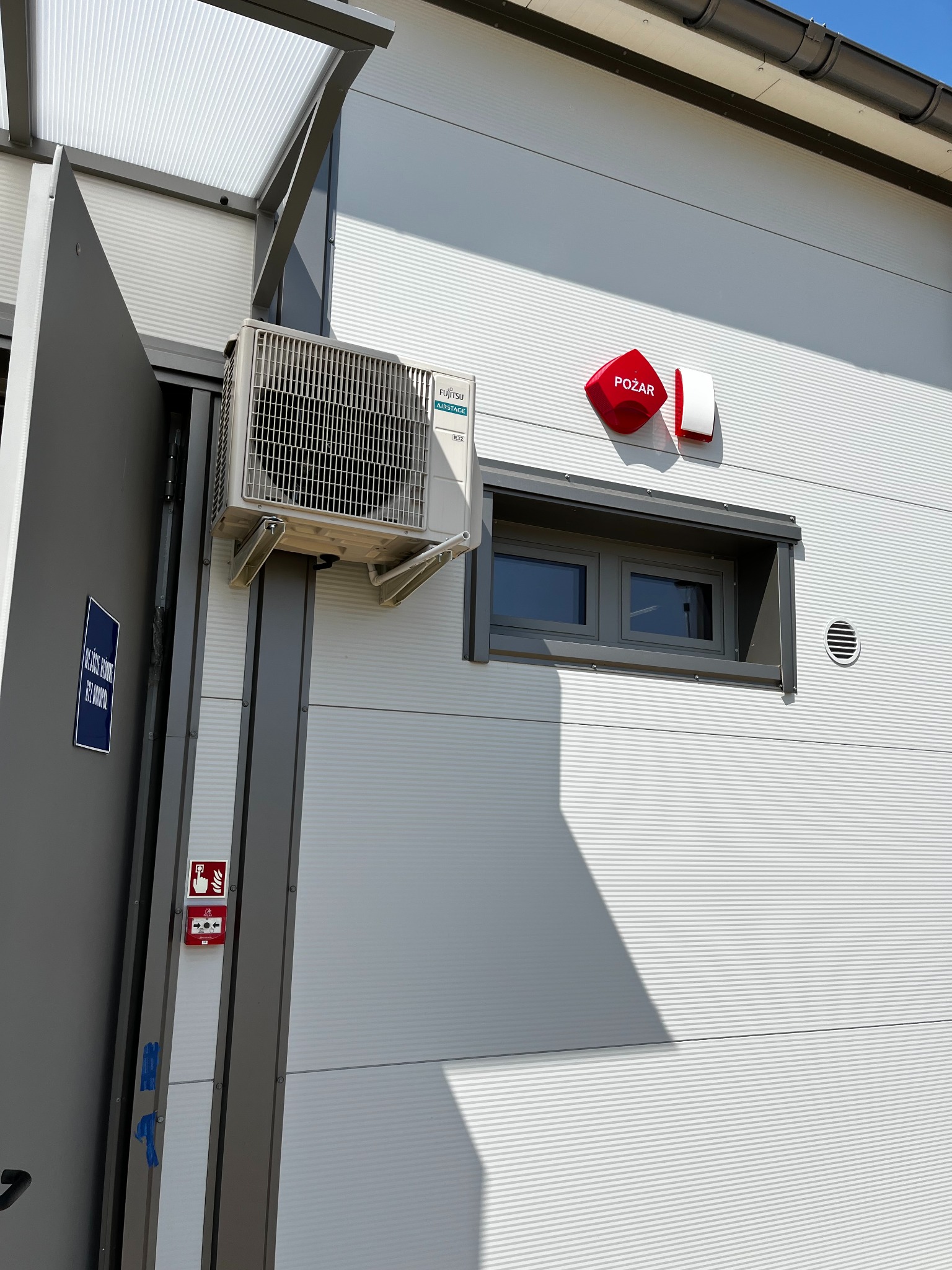 Biała elewacja budynku z zamontowanym alarmem przeciwpożarowym (czerwony klakson). Obok klimatyzator Fujitsu. Widoczne okno i tabliczka informacyjna na drzwiach.