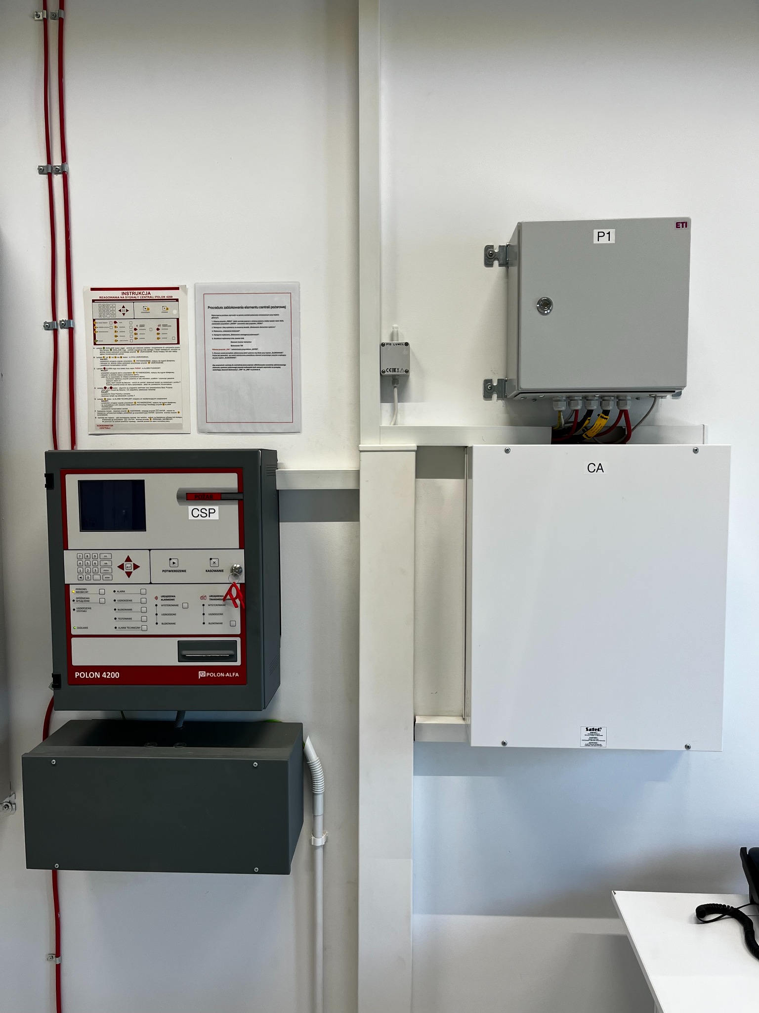 Centrala alarmowa Polon 4200 na ścianie, z instrukcją i schematem. Widoczne okablowanie i skrzynki elektryczne. Elementy systemu alarmowego wewnątrz budynku.