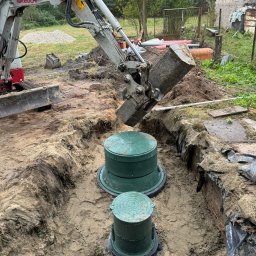 Agromar - Oczyszczalnie Przydomowe - Zbiorniki - Koparka umieszcza element betonowy w wykopie obok dwóch zielonych zbiorników z pokrywami, prawdopodobnie podczas montażu.