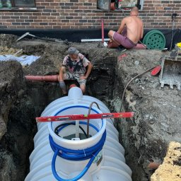 Agromar - Oczyszczalnie Przydomowe - Zbiorniki - Montaż białego zbiornika z niebieskimi rurami w wykopie obok budynku z czerwonej cegły, dwóch pracowników nadzoruje poziomowanie