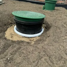 Oczyszczalnia przydomowa w trakcie instalacji, widoczny zielony właz i czarny zbiornik otoczony piaskiem, w tle budynek z czerwonej cegły.