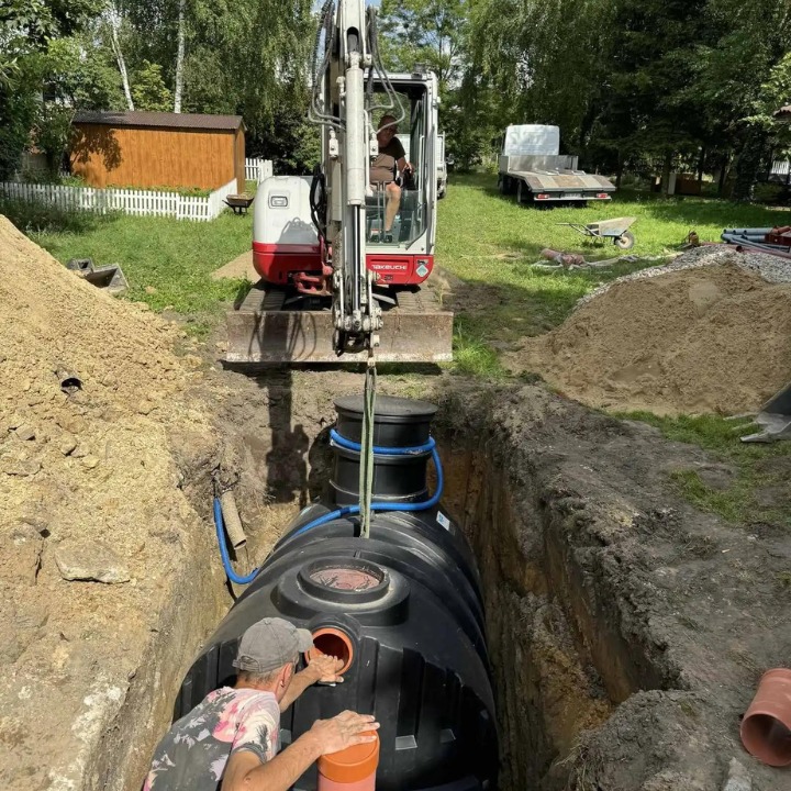 Montaż dużej, czarnej, plastikowej instalacji podziemnej w wykopie ziemnym przy użyciu minikoparki, widoczny pracownik w czapce, rury oraz sterta ziemi.