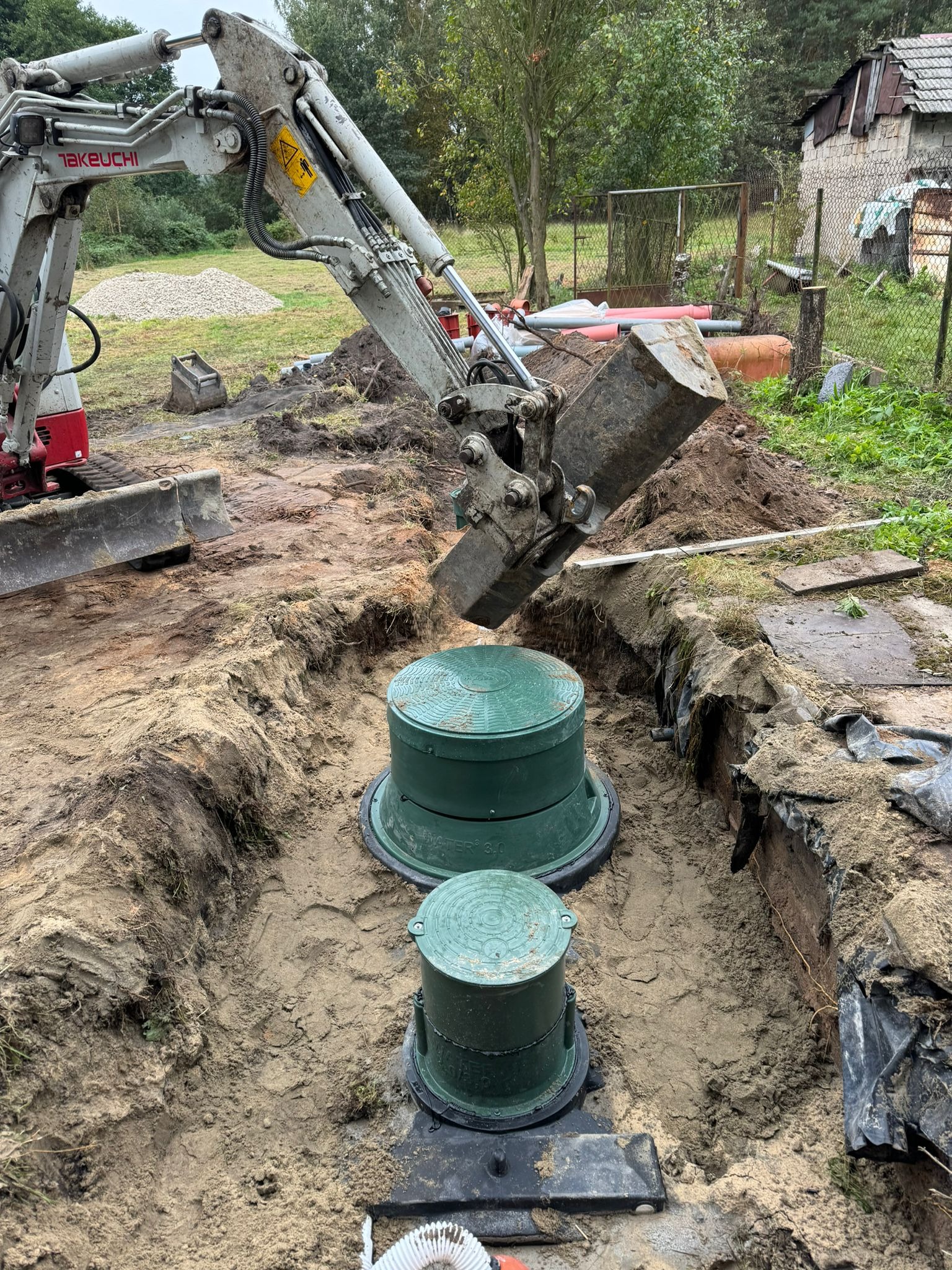 Koparka umieszcza element betonowy w wykopie obok dwóch zielonych zbiorników z pokrywami, prawdopodobnie podczas montażu.