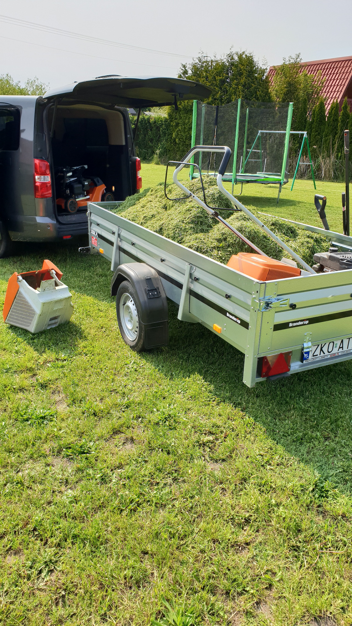 Przyczepa pełna skoszonej trawy, z kosiarką i pojemnikiem, zaparkowana obok auta z otwartym bagażnikiem na trawniku; w tle siatka trampoliny i dom.