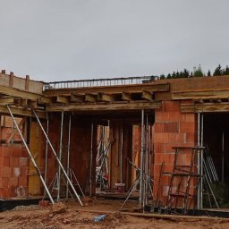 YN-BUD - Budowa domu w trakcie wznoszenia ścian z czerwonej cegły. Widoczne drewniane szalunki i metalowe podpory. Na ziemi leżą elementy konstrukcyjne.