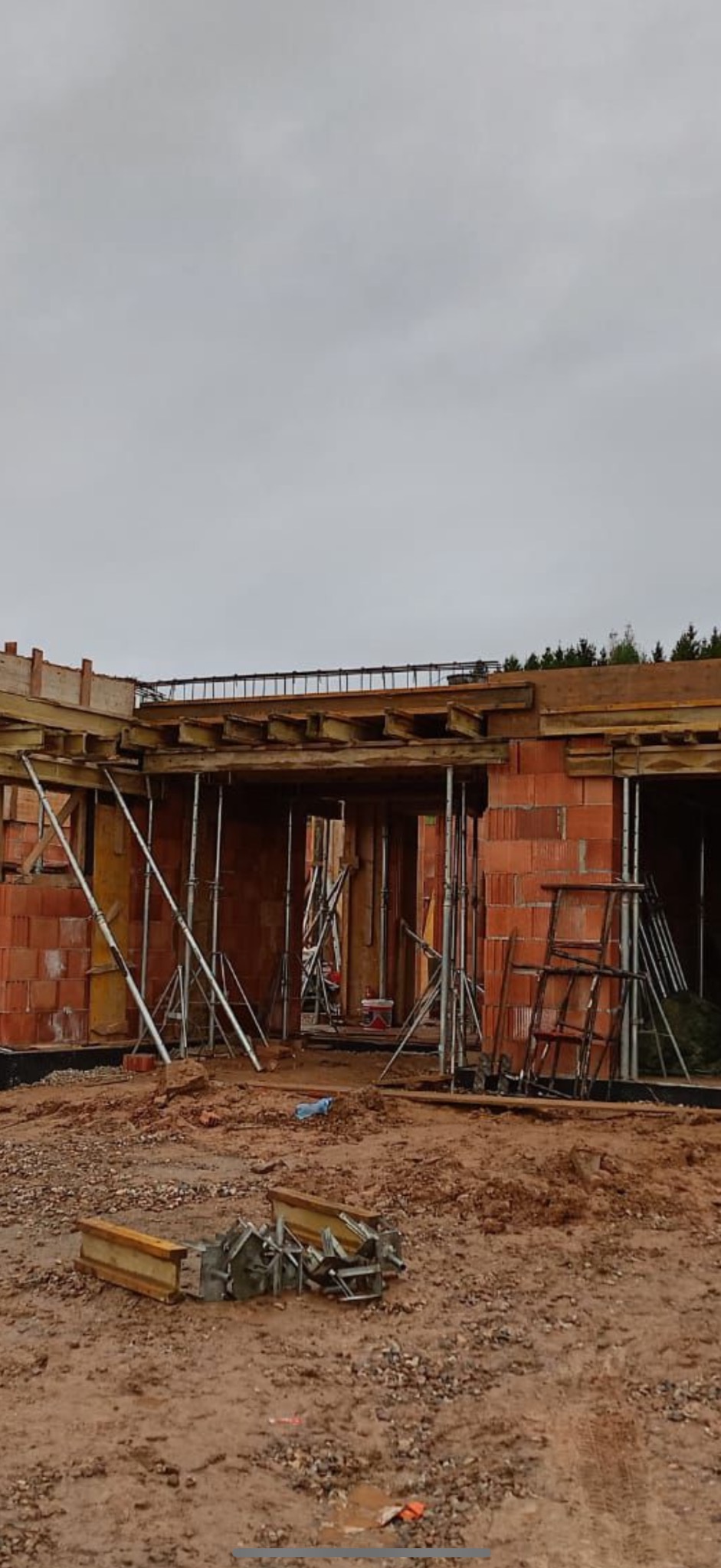 Budowa domu w trakcie wznoszenia ścian z czerwonej cegły. Widoczne drewniane szalunki i metalowe podpory. Na ziemi leżą elementy konstrukcyjne.