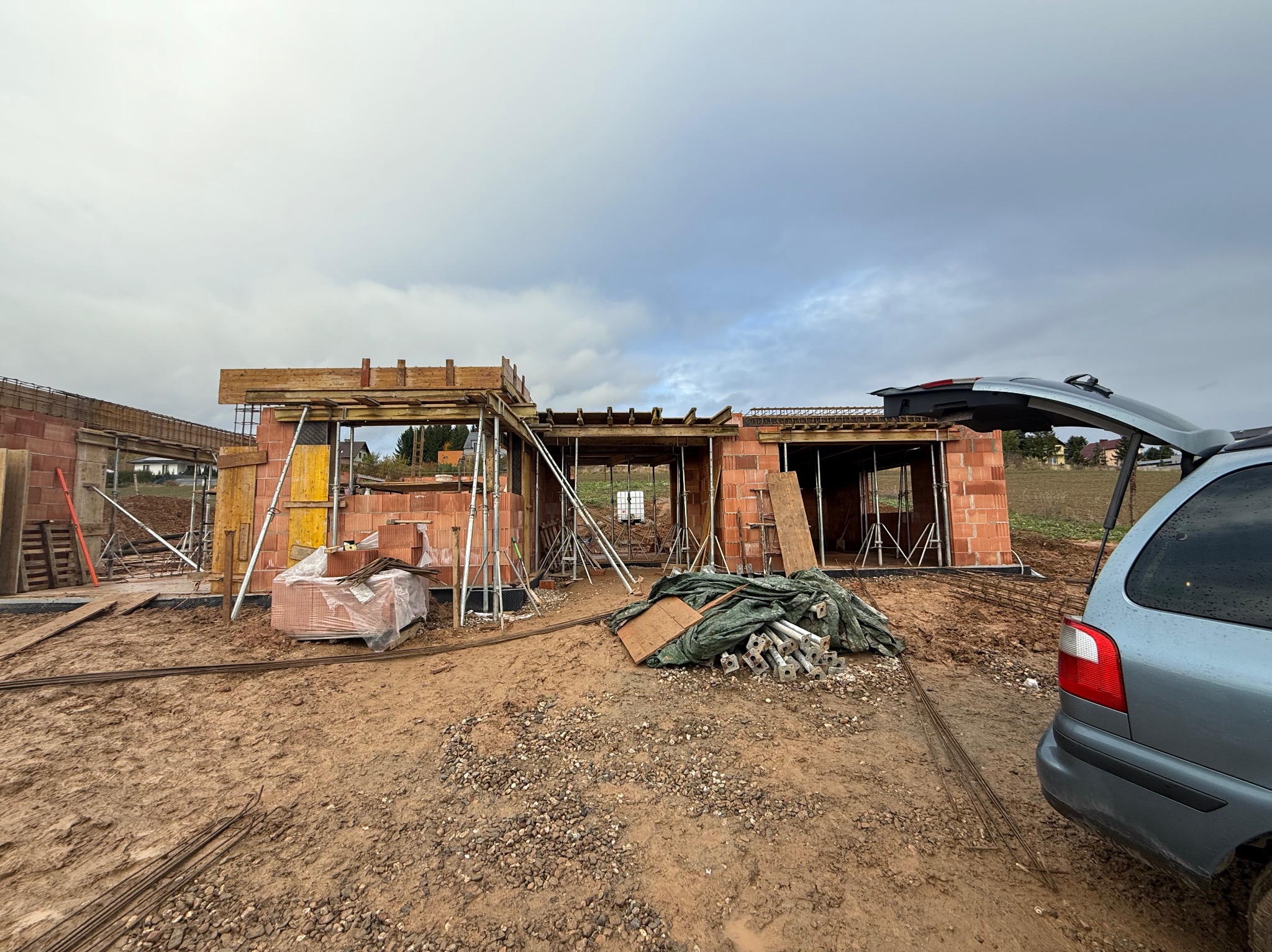 Budowa domu w Jeleniej Górze w trakcie wznoszenia ścian z czerwonej cegły. Widoczne podpory, szalunki i materiały budowlane. Otwarty bagażnik samochodu w tle.