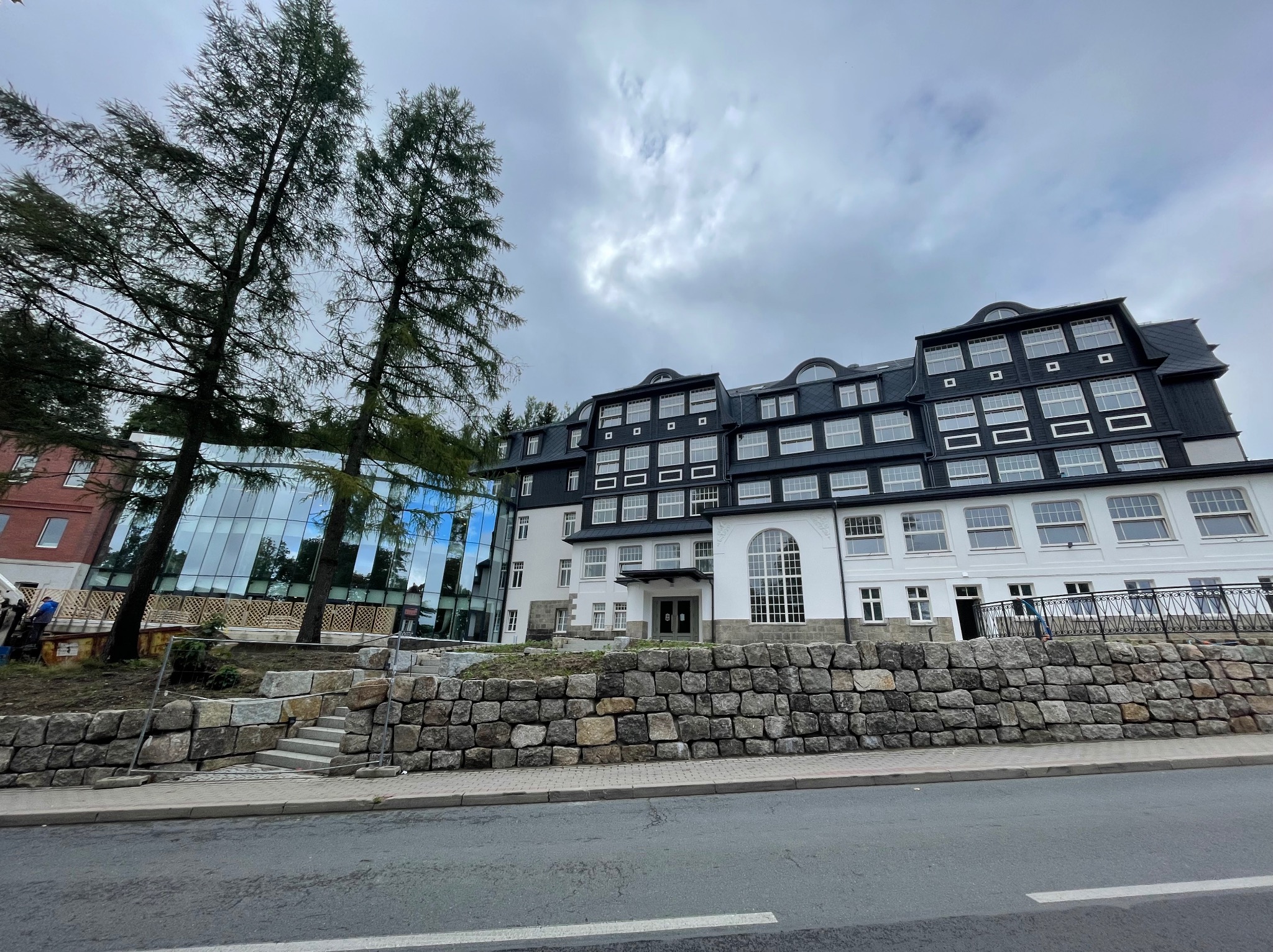 Elewacja budynku z czarnym dachem i białymi ścianami, widoczna z poziomu ulicy. Kamienny murek i drzewa w tle. Pochmurne niebo.