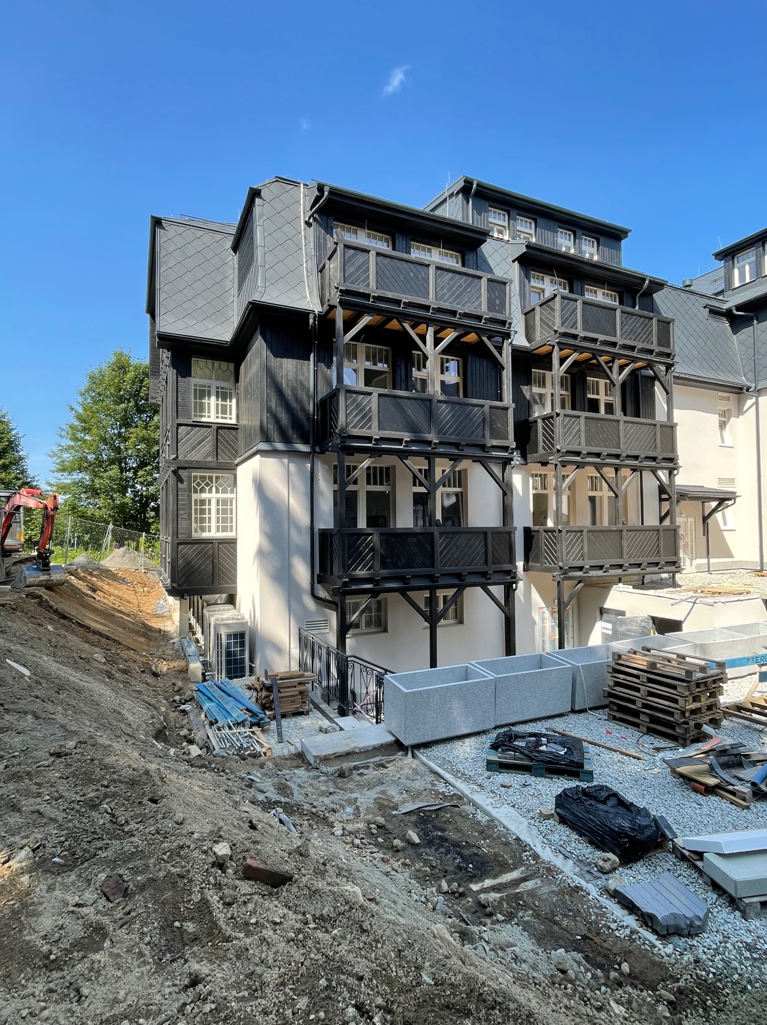 Nowoczesny budynek z balkonami w stylu góralskim w trakcie prac wykończeniowych. Widoczny wykop, materiały budowlane i minikoparka. Słoneczny dzień.