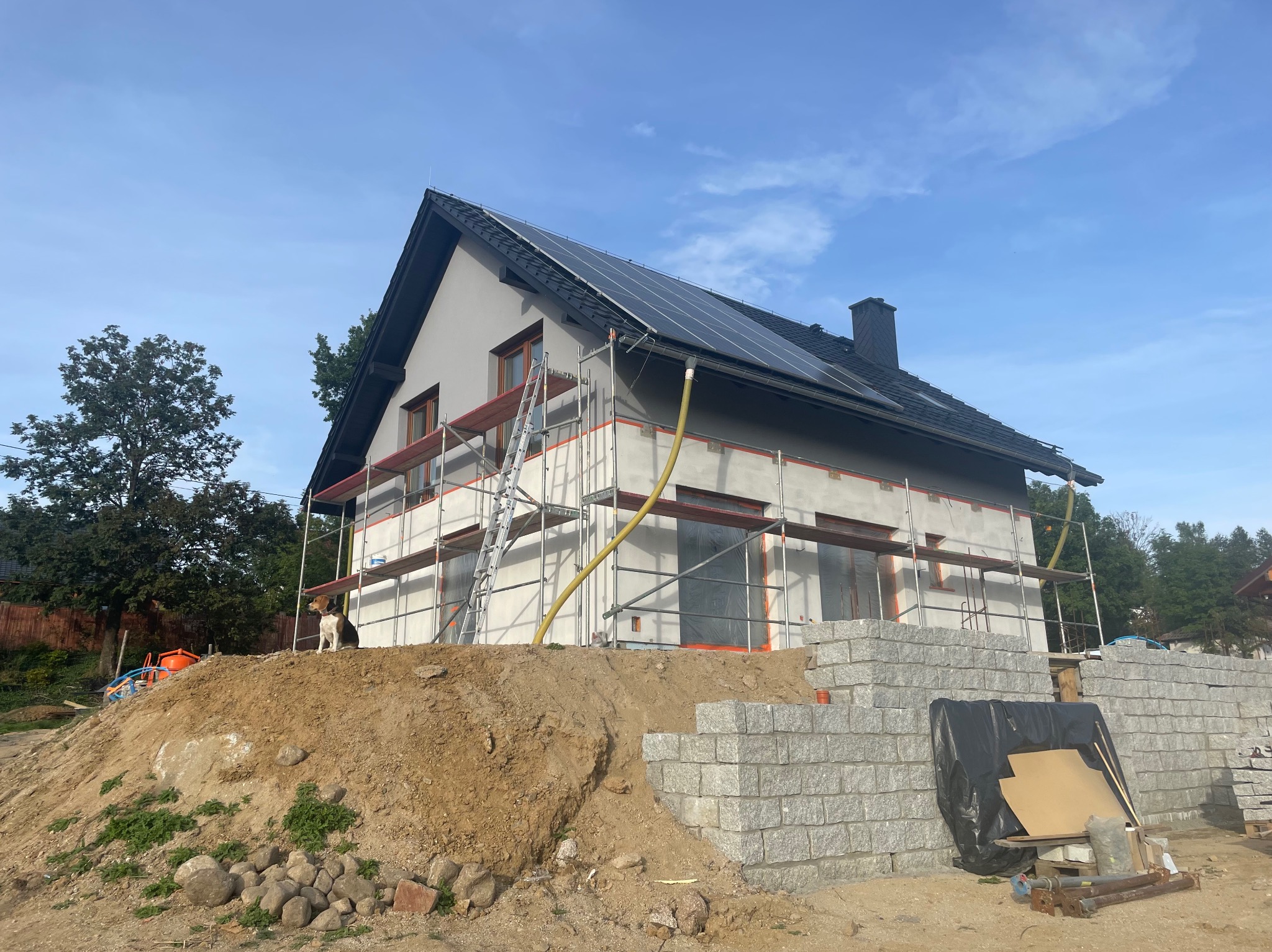Dom w trakcie ocieplania, z panelami słonecznymi na dachu, otoczony rusztowaniem. Na pierwszym planie pies siedzący na usypanym wzgórzu ziemi.