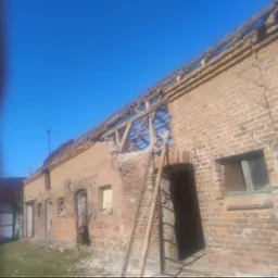 Ceglany budynek w trakcie rozbi&oacute;rki, z widoczną drewnianą konstrukcją dachu i drabiną opartą o ścianę. Fragmenty ściany usunięte, gruz na ziemi.