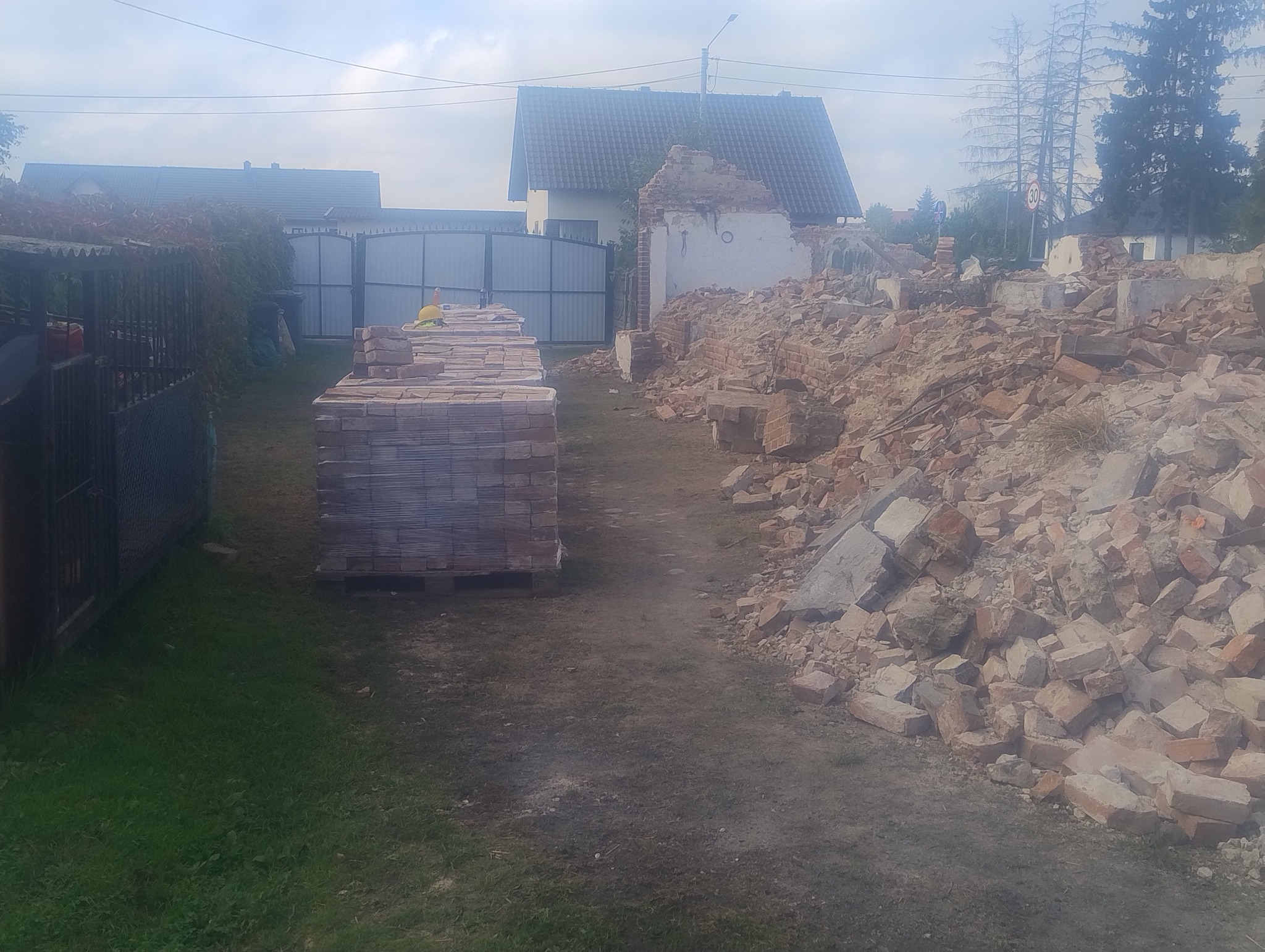Stos cegieł obok gruzowiska po rozbiórce budynku. Widoczny fragment ściany i ogrodzenie w tle. Prace rozbiórkowe w toku.