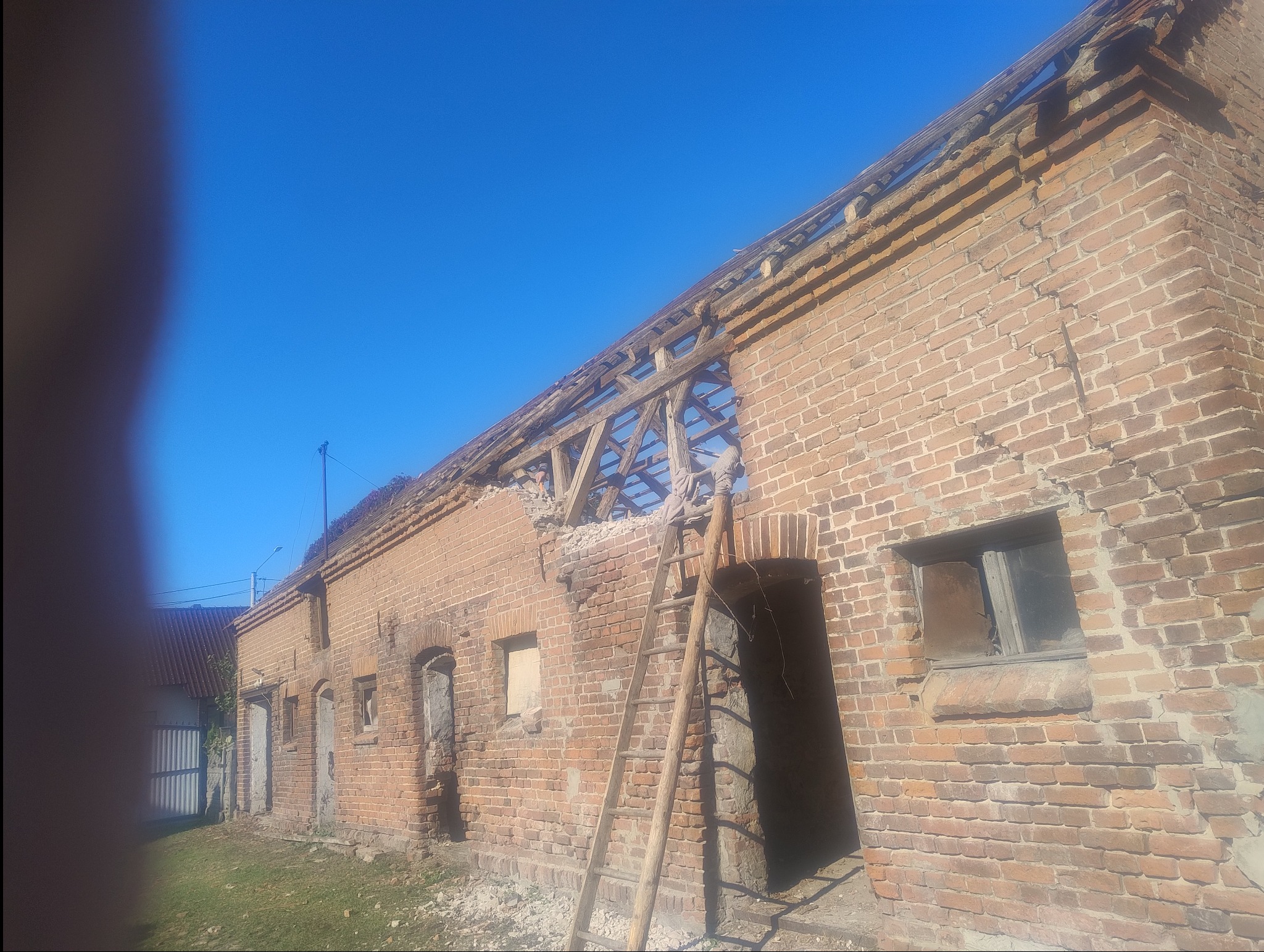 Ceglany budynek w trakcie rozbiórki, z widoczną drewnianą konstrukcją dachu i drabiną opartą o ścianę. Fragmenty ściany usunięte, gruz na ziemi.