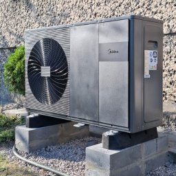 Hali Invest Kamil Halibożek - Zewnętrzna jednostka pompy ciepła Midea, zamontowana na betonowych bloczkach fundamentowych, z widocznym przewodem instalacyjnym przy ziemi, na tle chropowatej elewacji budynku z oknem.