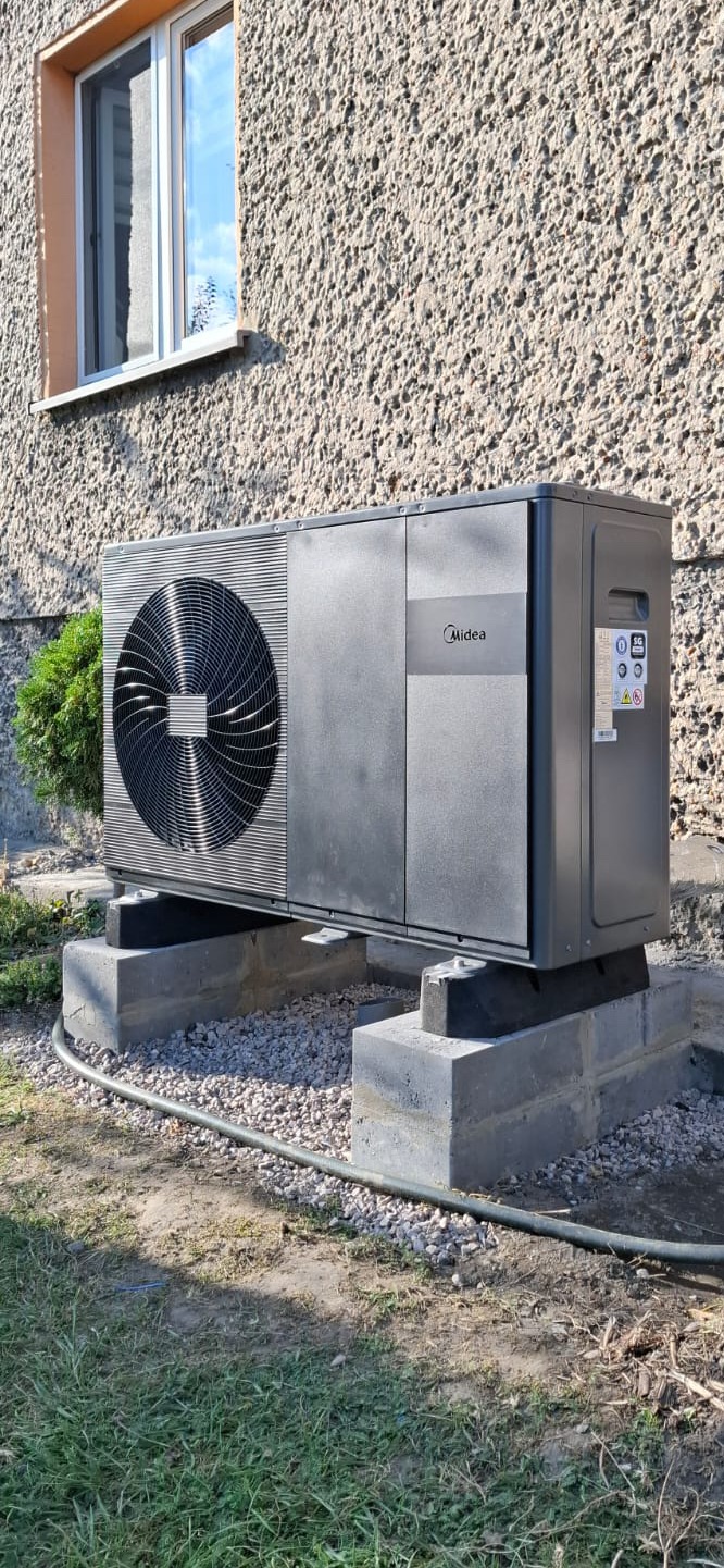Zewnętrzna jednostka pompy ciepła Midea, zamontowana na betonowych bloczkach fundamentowych, z widocznym przewodem instalacyjnym przy ziemi, na tle chropowatej elewacji budynku z oknem.