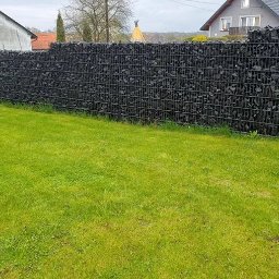 KRAK FENCE SPÓŁKA Z OGRANICZONĄ ODPOWIEDZIALNOŚCIĄ - Gabionowe ogrodzenie z czarnym wypełnieniem kamiennym, widok z poziomu trawnika, w tle domy jednorodzinne.