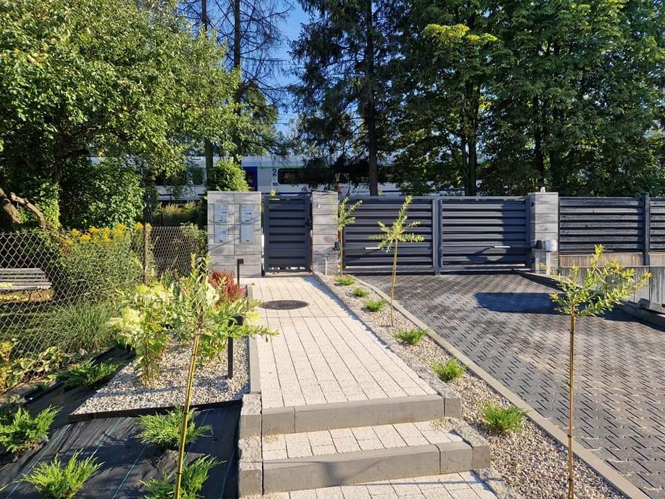 Nowoczesna posesja z grafitową bramą przesuwną i ogrodzeniem z poziomych paneli, kamienna ścieżka prowadząca do wejścia, młode drzewka wzdłuż podjazdu, widok na zadbany ogród i budynek w tle.