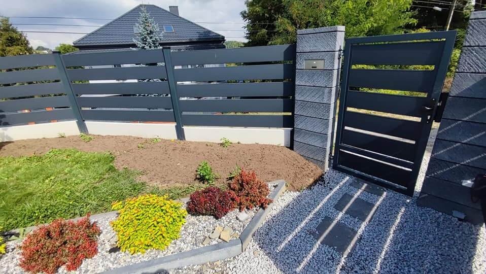 Nowoczesne ogrodzenie z poziomych, szarych paneli i betonowych słupków z zamontowaną skrzynką na listy, dopasowana kolorystycznie brama z poziomymi szczelinami, świeżo zagospodarowany teren...