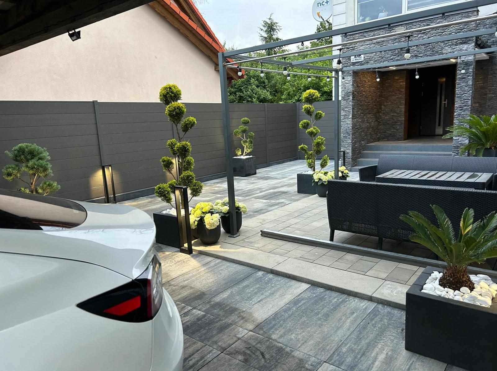 Nowoczesny taras z szarym ogrodzeniem panelowym, oświetleniem LED, drzewkami bonsai w donicach i widocznym fragmentem białego samochodu Tesla.