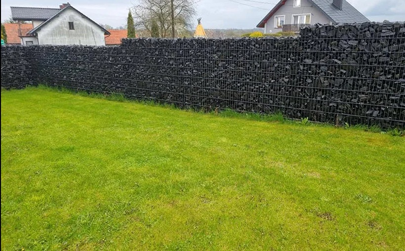 Gabionowe ogrodzenie z czarnym wypełnieniem kamiennym, widok z poziomu trawnika, w tle domy jednorodzinne.