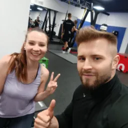 Selfie w siłowni: uśmiechnięta kobieta pokazuje znak 'V', mężczyzna kciuk w górę; w tle sprzęt do treningu TRX i inne osoby ćwiczące.