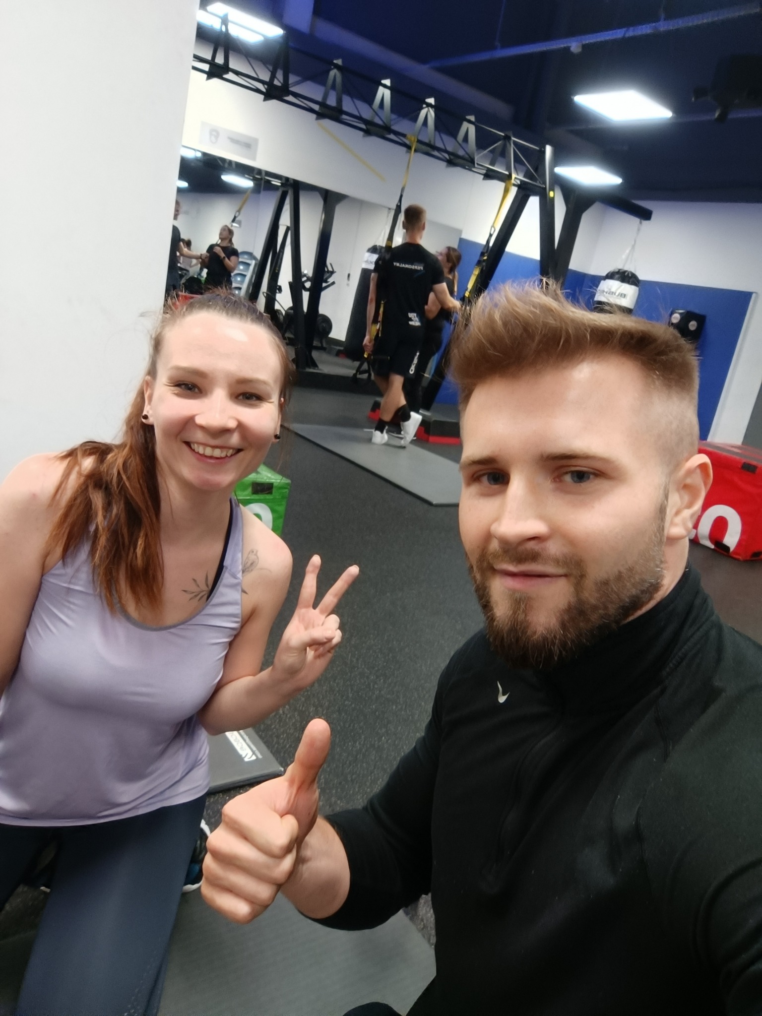 Selfie w siłowni: uśmiechnięta kobieta pokazuje znak 'V', mężczyzna kciuk w górę; w tle sprzęt do treningu TRX i inne osoby ćwiczące.