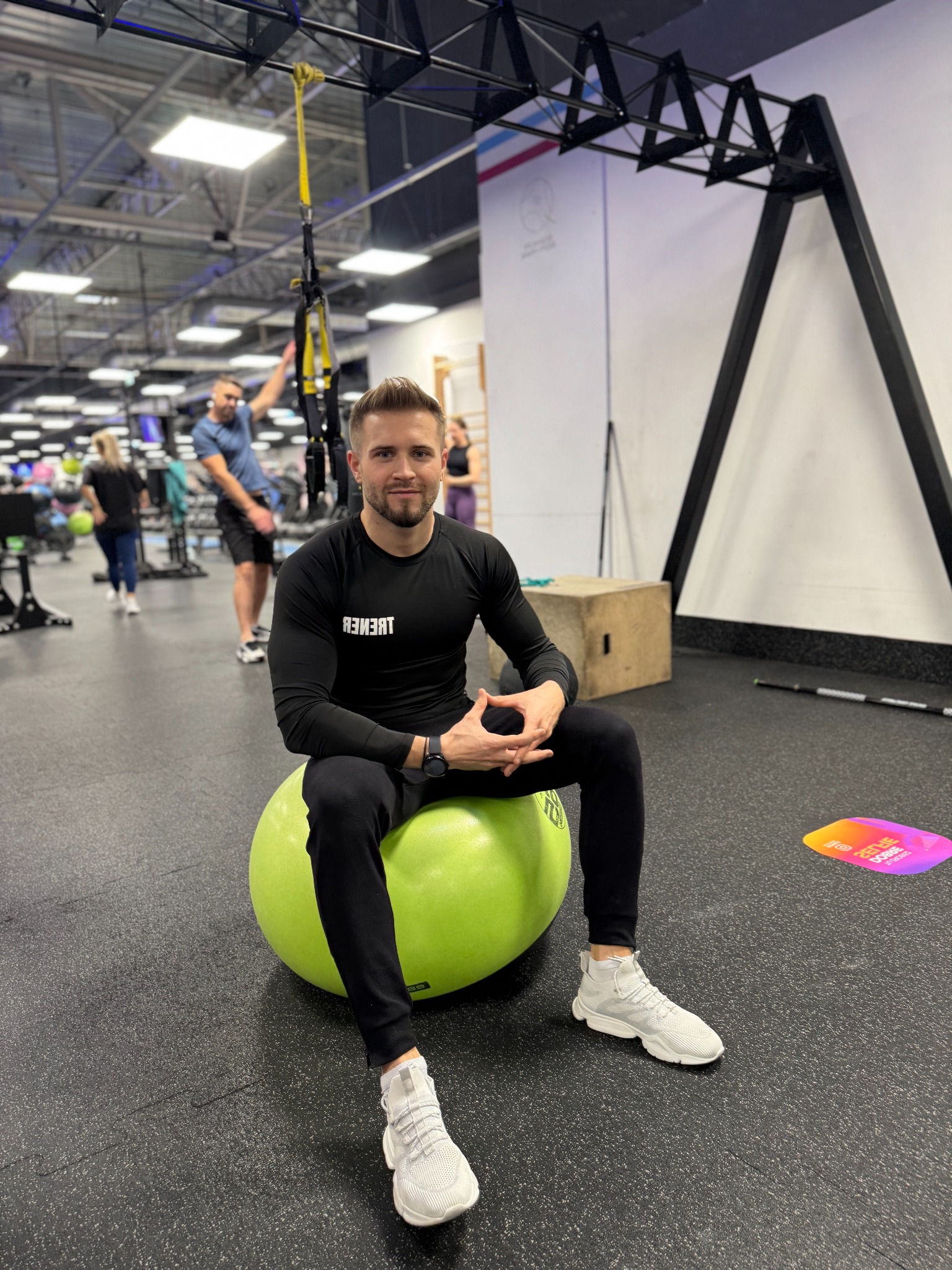 Uśmiechnięty trener personalny siedzi na zielonej piłce gimnastycznej w siłowni, w tle widoczni ćwiczący i sprzęt do ćwiczeń.