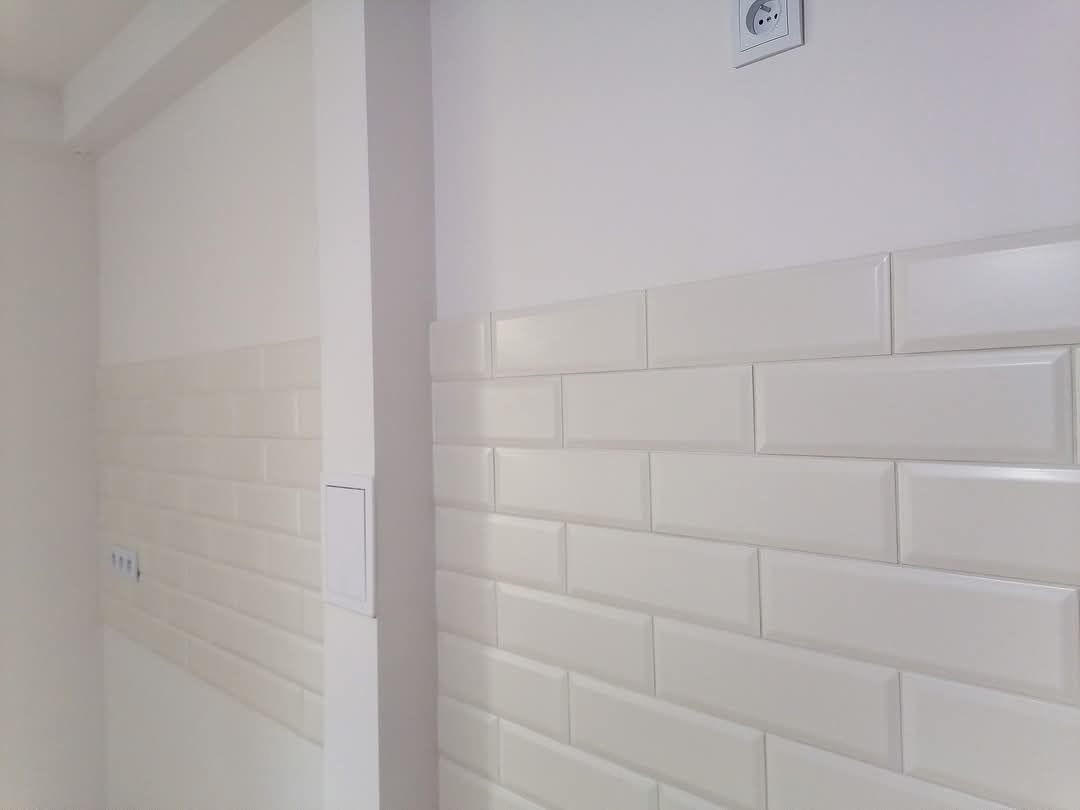 Ściana wyłożona białymi, prostokątnymi płytkami ceramicznymi typu metro, z widocznym gniazdkiem elektrycznym powyżej, w minimalistycznym wnętrzu.