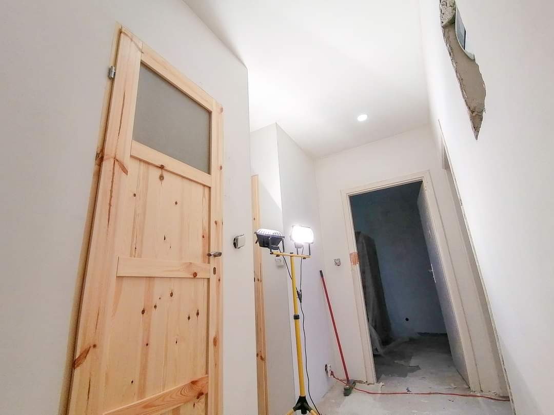 Montaż drewnianych drzwi z przeszkleniem w trakcie prac wykończeniowych, widoczny reflektor budowlany i fragment uszkodzonej ściany.