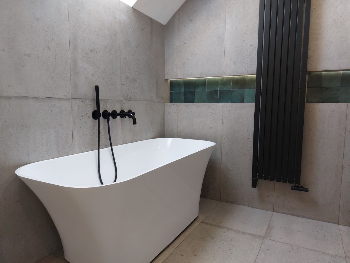 Nowoczesna łazienka z wolnostojącą białą wanną, czarną armaturą i grzejnikiem drabinkowym na tle szarych płytek imitujących beton, akcentowana paskiem zielonych płytek.