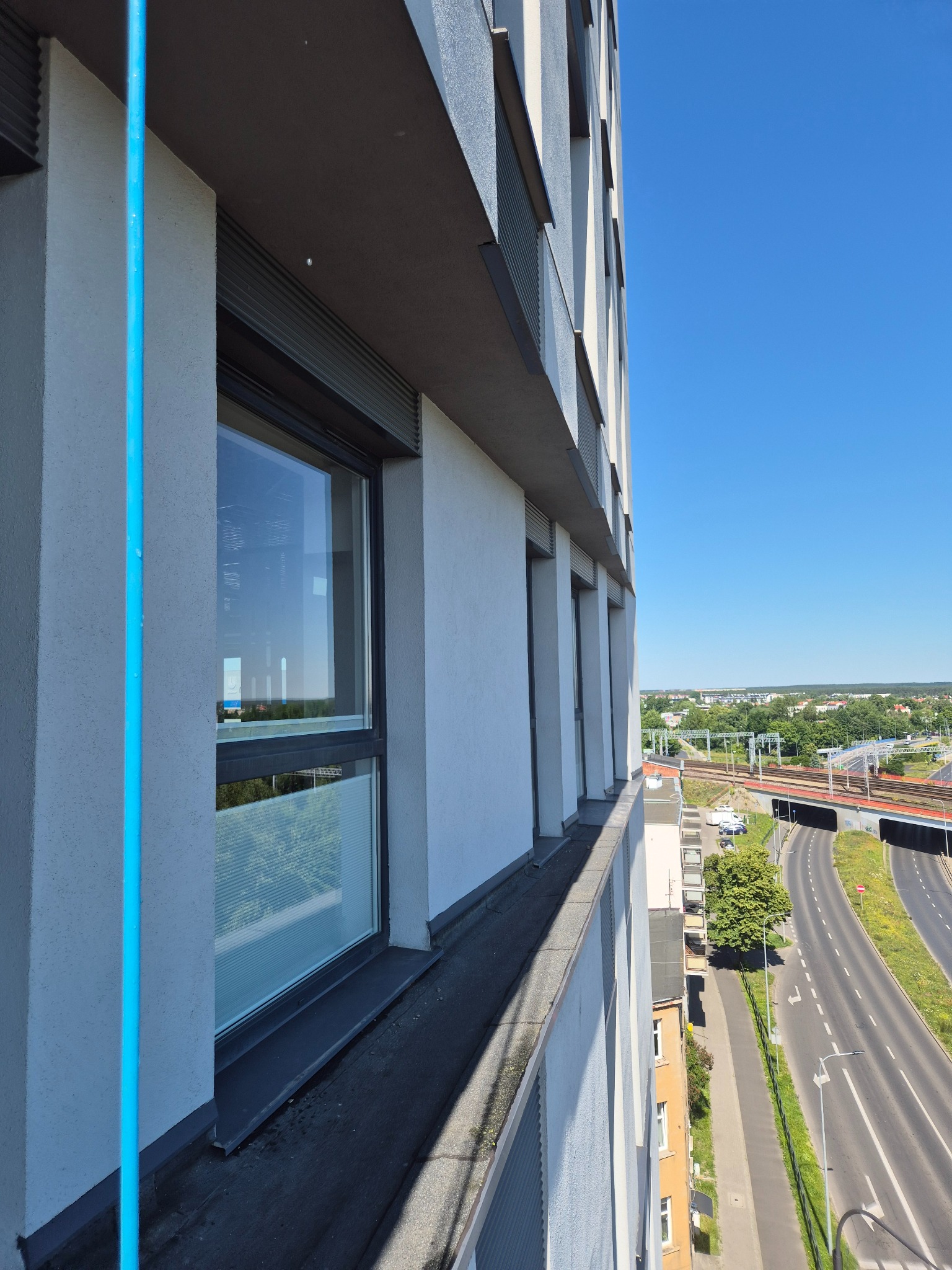 Widok z wysokości na okna budynku z aluminiowymi roletami, błękitny przewód na elewacji, w tle panorama miasta i autostrada w słoneczny dzień.