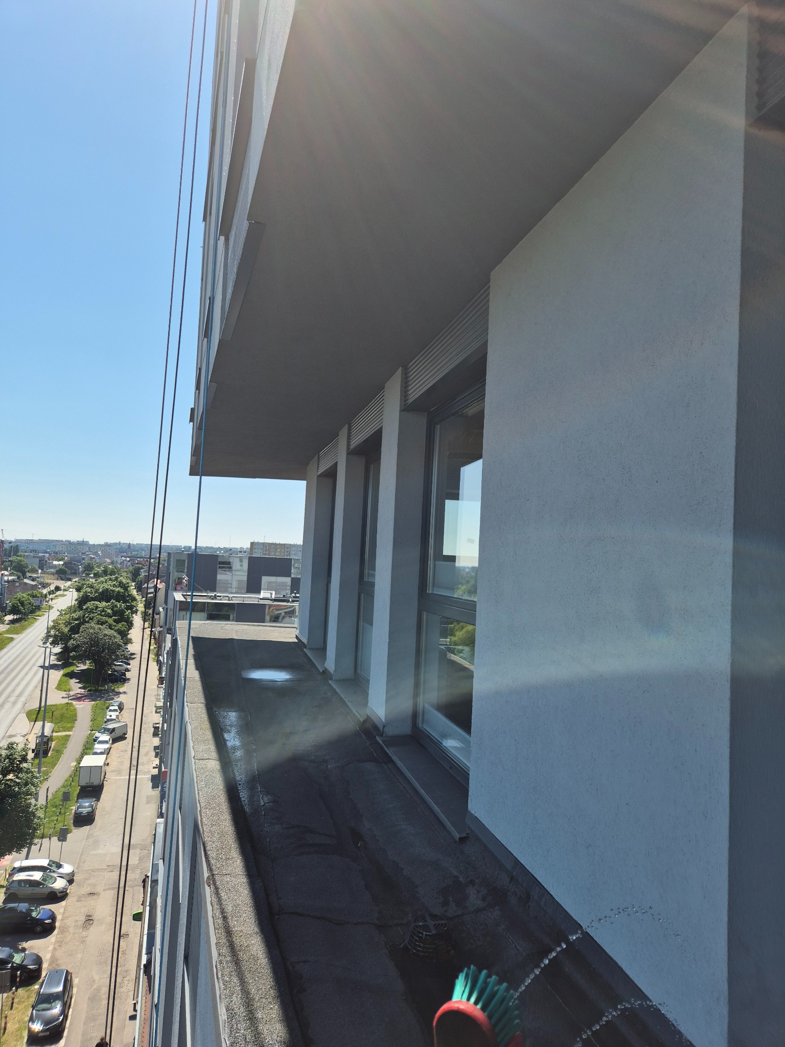 Mycie okien na wysokości w Witkowie. Widok z góry na balkon wieżowca podczas czyszczenia elewacji i okien za pomocą szczotki na kiju. W tle panorama miasta.