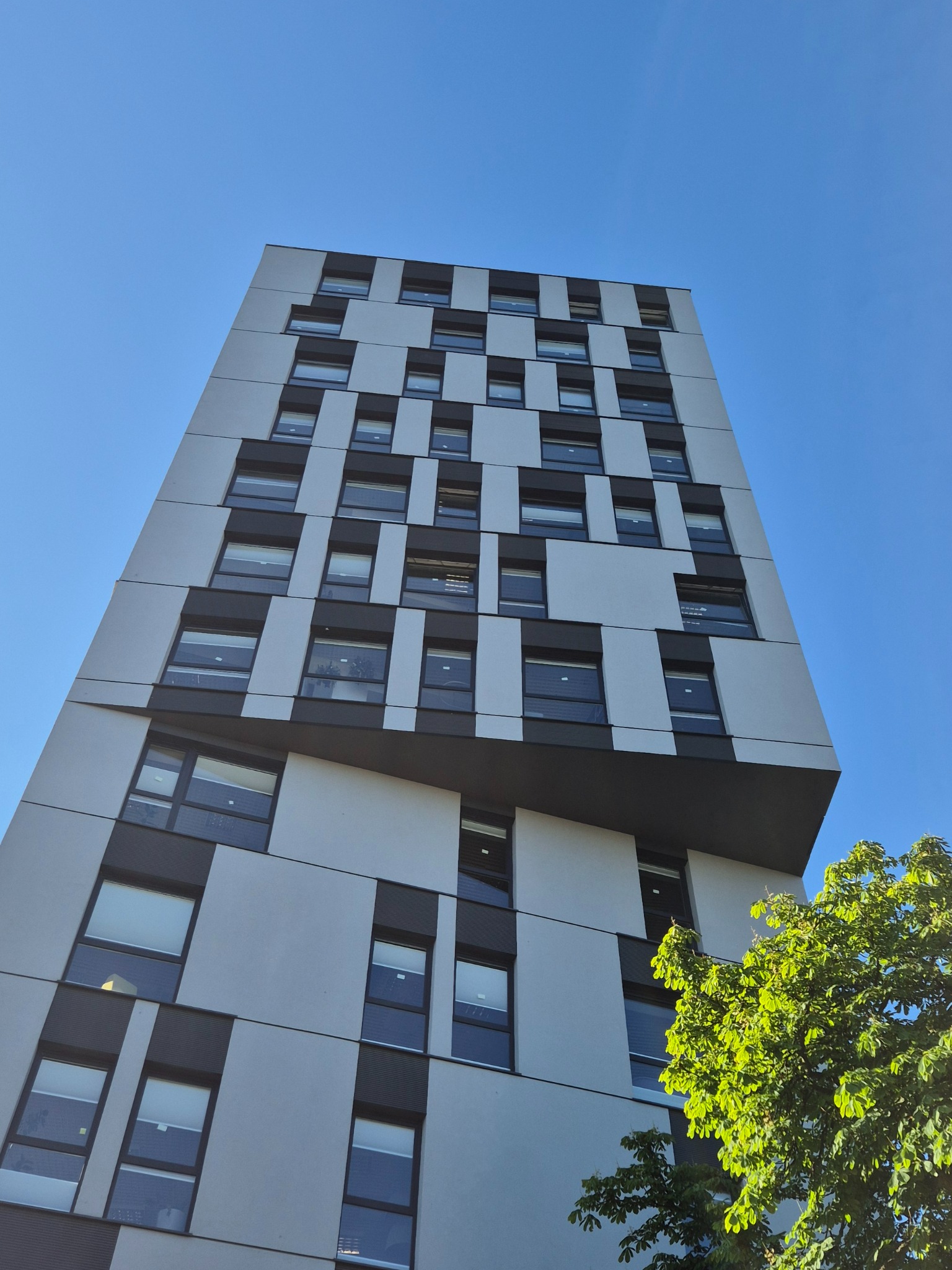 Wysoki budynek biurowy z licznymi oknami i nowoczesną fasadą w biało-szarej kolorystyce, widziany z dołu na tle błękitnego nieba, z fragmentem zielonego drzewa w rogu.