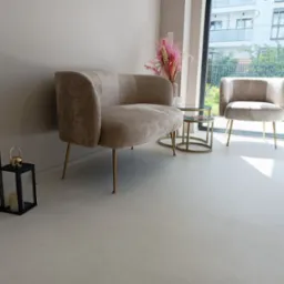 Jasnoszara, gładka posadzka w pomieszczeniu z beżową sofą i fotelem na złotych nóżkach, stolikami kawowymi i dekoracyjnymi lampionami; widok na zewnątrz przez duże okno.